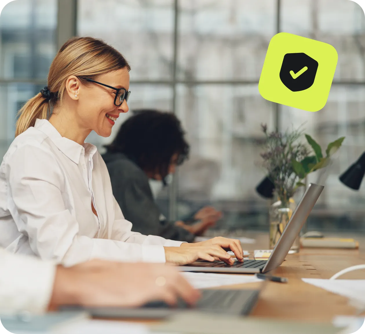 Woman smiling while working on a laptop in an office, with a security checkmark icon above.