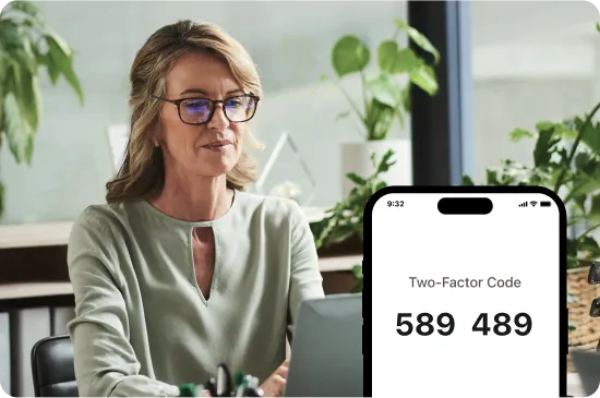 Woman at a desk using a laptop, with a phone screen showing a two-factor authentication code.